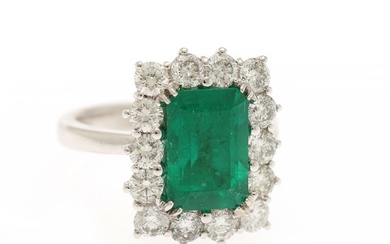 An emerald and diamond ring set with an emerald-cut emerald encircled by numerous brilliant-cut diamonds, mounted in 18k white gold. Size 55.5.