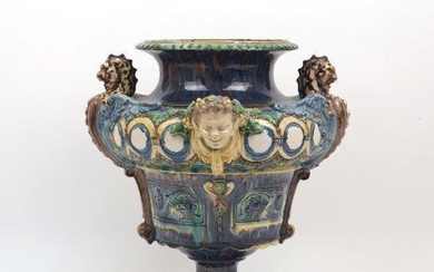 A Continental majolica urn, 19th century, of blue-brown slip glaze with green and yellow highlights, the handles in the form of dragons, applied bacchic masks and panels with sea serpents to the body, modelled on socle base, 35.6cm high