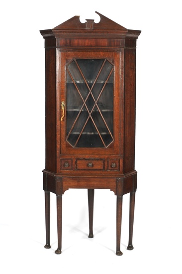 A Georgian glazed oak corner cupboard. With a broken swan neck pediment above moulded cornice, the glazed cupboard door with X-shaped astragals enclosing three shaped shelves, above a narrow frieze drawer and two dummy door fronts, on canted base with...