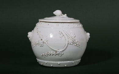 A CHINESE BLANC-DE-CHINE 'PRUNUS' JAR AND COVER. Qing Dynasty, 18th Century. Formed as a barrel with a pair of lion head handles and a blooming prunus applied to one side, with bands of studs to the rims, the flat cover with a peach finial, 13cm H...