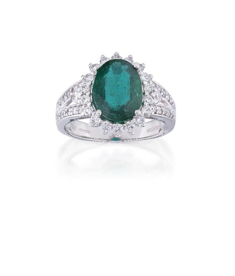 An emerald and diamond ring, the oval emerald, weighing approximately 2.82 carats, to a brilliant-cut diamond surround and bifurcated shoulders, mounted in 18ct white gold, Birmingham hallmarks, maker's mark DJM for David Jerome, ring size M...