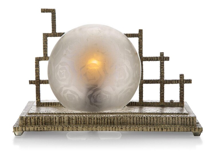 An Art Deco metal table lamp, c.1930, The stepped metal base cast with floral motifs and ribbed edges, the globular frosted shade held by openwork gates, two holes at one end, 16.5 cm high, 24.5 cm long