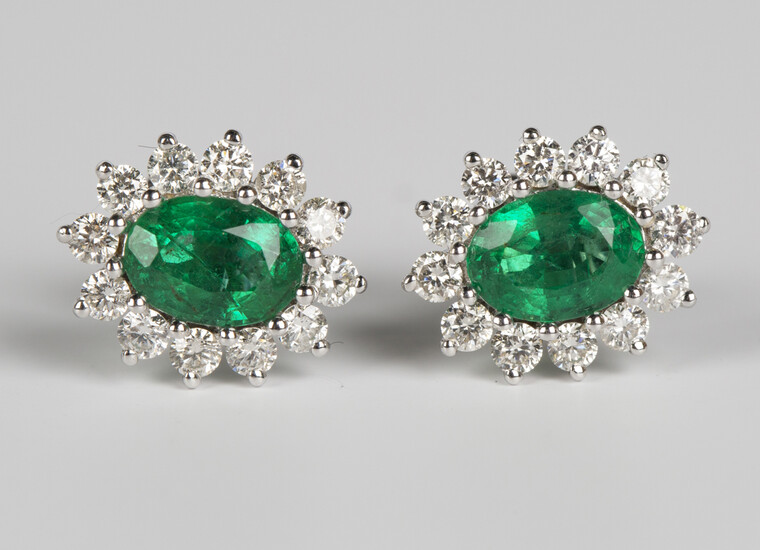A pair of 18ct white gold, emerald and diamond earrings, each claw set with an oval cut emerald with