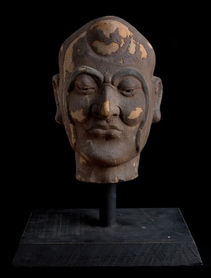 A Chinese clay head of a Luohan, Yuan/Ming dynasty, modelled and carved of a full rounded head with long sinuating eyebrows extending to his jawline, downcast eyes above a rounded nose, flanked by long pendulous earlobes, with a wood stand, 31cm...
