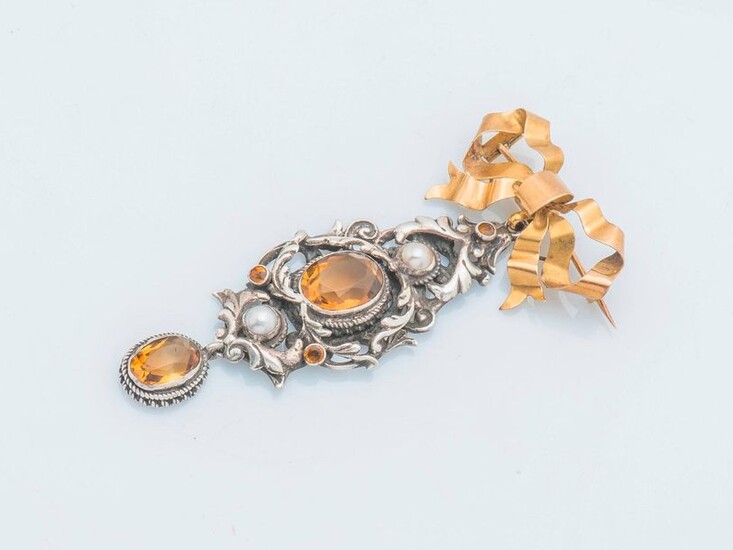 Brooch in 18-carat yellow gold (750 thousandths) and silver (800 thousandths) adorned with a ribboned bow, holding a stylised element of intertwined acanthus leaves, centred on an orange stone enhanced with two pearls, holding an oval-cut orange stone...