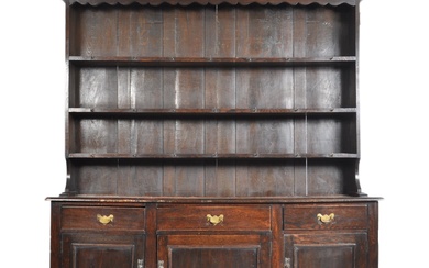 An 18th century George III oak Welsh dresser. Raised on bracket feet supporting a wide body of three short drawers over cupboards beneath. Planked top with rounded leading edges. Above, an upright gallery plate rack with suspended pediment. Dismantles...