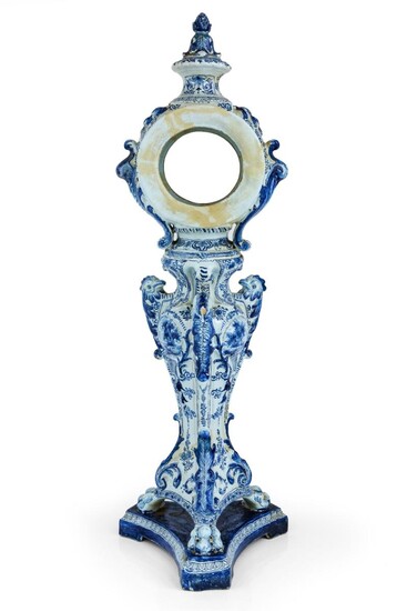 A Dutch Delft blue and white clock-case, 19th century, pseudo blue Adrianus Kocks AK monogram / 99 mark, the cylindrical case surmounted by a foliate finial, the baluster stem painted with three landscape vignettes, one with a farm girl, divided by...