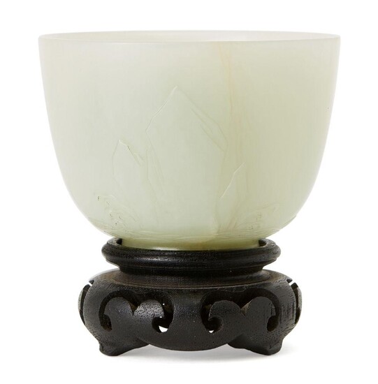 A Chinese celadon jade cup, late Qing dynasty, finely carved in low relief with two rock formations arising from crashing waves, slightly flared short foot, 5.5cm high, later wood stand