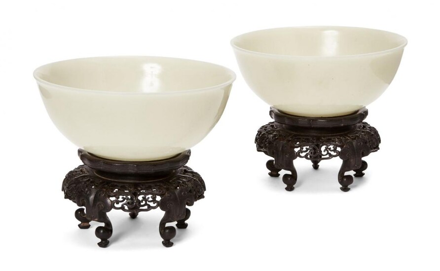 A pair of Chinese pale celadon jade bowls, 19th century, each carved with deep sides and a slightly everted mouth rim, base with an incised apocryphal Qianlong mark, 17.5 cm diameter wide, both on finely carved openwork hardwood stands