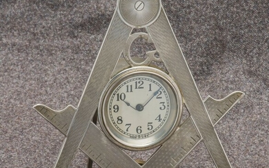 Pendulette en métal argenté formant le compas et l’équerre surmonté du G. Le cadran au centre argenté, à chiffres arabes et chemin de fer. Mouvement mécanique (ne fonctionne pas). H. 16 cm