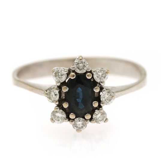 A sapphire and diamond cluster ring set with an oval-cut sapphire encircled by numerous brilliant-cut diamonds, mounted in 14k white gold. Size 55.5.