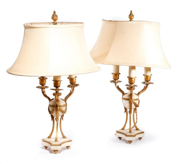 A PAIR OF FRENCH ORMOLU AND WHITE MARBLE TABLE LAMPS IN LOUIS XVI STYLE LATE 19TH CENTURY in the form of atheniennes, with three lights, with fluted urn nozzles and petal drip-pans, on hoof feet and a triform base, with shades and wired for...