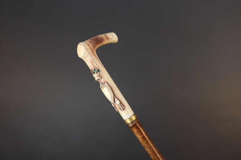 Cane with square knob. Bamboo shaft. Carved antler grip decorated with a coat of arms surmounted by a crown decorated with small green cabochons (two missing). Height 90,5 cm. Crack in the pommel