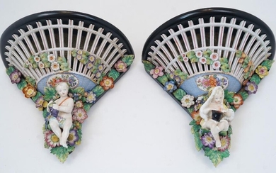A pair of German porcelain wall brackets, 19th century, of pierced trellis design with encrusted flowers terminating in putti, each with ebonised wood wall mounts, marks obscured, 25cm high, 30cm wide, 15.5cm deep (2)