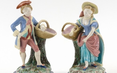 PAIR OF ROYAL WORCESTER MAJOLICA FIGURES A boy and a girl in 18th Century costume, both carrying a basket resting on a tree stump. I...