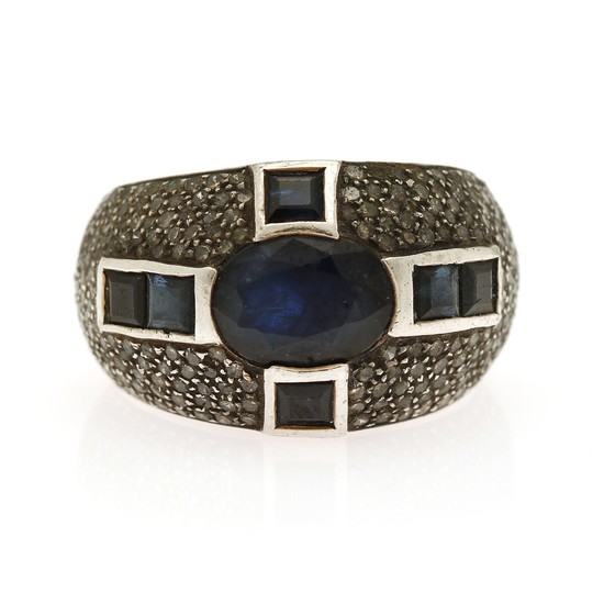 A sapphire and diamond ring set with an oval- and six square-cut sapphires encircled by numerous single-cut diamonds, mounted in silver. Size 58.
