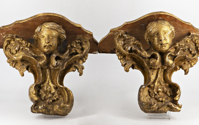 Coppia di mensole in legno intagliato e dorato a forma di testa d'angelo su volute, secolo XVIII. Piani sagomati e…