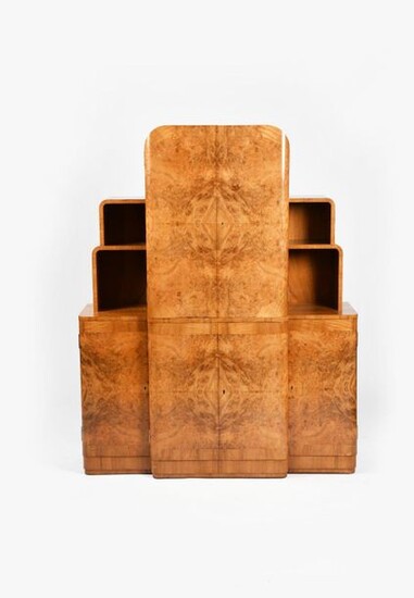 An Art Deco burr walnut veneer cocktail cabinet probably Epstein, L'Odeon form, two central twin door fitted cabinets, the top compartment with electric light, flanked with smaller side cabinets below stepped shelves, fitted with suite of glasses...