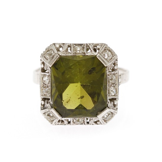 An Art Deco peridote and diamond ring set with a fancy-cut peridote encircled by numerous old-cut diamonds, mounted in platinum. Size 58.