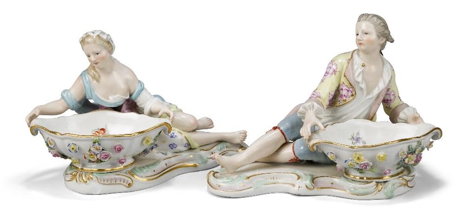A pair of Meissen figural sweetmeat dishes, late 19th/early 20th century, blue crossed sword marks, incised numerals 2872, 2875 and 51, modelled as a finely dressed male and female reclining holding flower-encrusted bowls decorated with flowers and...