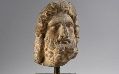 Head from a statue probably representing the god Zeus or Jupiter. He wears a beard and a hairstyle composed of long wavy locks encircled by a ribbon. The back part of the hair is distributed on either side of a median parting. The wide-open eyes and...