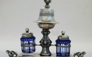 A Continental white metal inkstand, with bell on stand to centre with elaborate scrolling knopped stem flanked by a blue and clear glass pounce pot and matching inkwell with white metal covers, on shaped base with scrolling foliate handles, stamped...