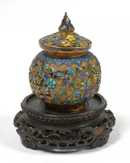 Small round box covered in gilded fishnet decorated with enamelled silver floral decoration.Chinese work. Circa 1900. Resting on a carved wood base. (Light blows). H.:+/-8,2cm.