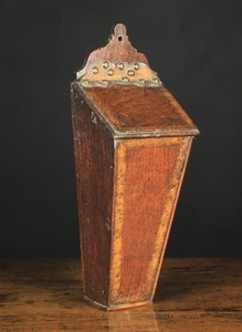A 19th Century Boarded Oak Candle-box inlaid with burr figured bands to the borders. The sloped lid hinged by a studded leather strap, above a tapering box with shaped back board pierced with hanging hole, 17½ in (44 cm) high, 6 in (15 cm) wide, 6