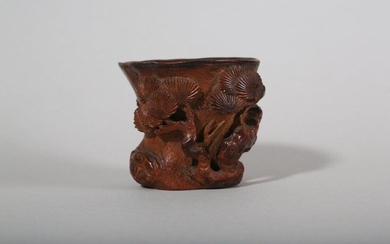 A CHINESE CARVED BAMBOO 'PINE TREE' LIBATION CUP. 17th Century. Naturalistically carved as a burled tree trunk enveloped with gnarled branches meandering around the vessel, the branches depicted bearing clusters of large pine leaves, the interior...
