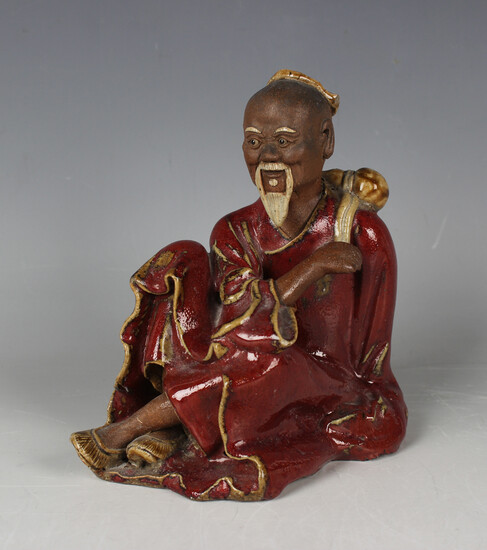 A Chinese sang-de-boeuf glazed stoneware figure of a seated sage, modelled with gourd water bottle s