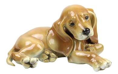 Allach Lying dachshund puppy, designed by Theodor Kärner (Hohenberg a. d. Eger 1884-1966 Munich), manufactured by Porzellanmanufaktur Allach, slightly cream-coloured porcelain, naturalistically painted under glaze, L 17 cm, H 9.5 cm, with green...