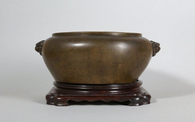 A CHINESE BRONZE INCENSE BURNER. Qing Dynasty. The gently tapered body cast to the rounded shoulder with two lion head handles, the surface with an even warm patina, the base with a six-character Xuande mark, together with a wood stand, 23.5cm...