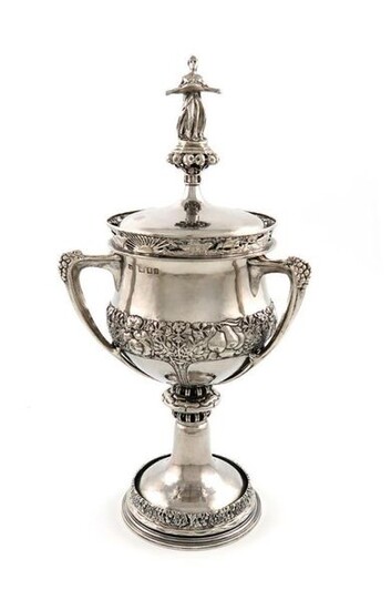 By Alwyn Carr, an Arts and Crafts silver three-handled cup and cover, London 1921, the circular bellied body spot-hammered and with chased foliate and fruit decoration, with three scroll handles mounted with three bunches of grapes, on a knopped stem...