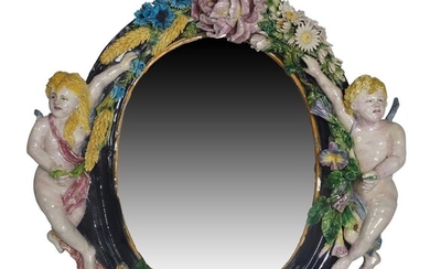 A Continental majolica style mirror, early 20th century, with oval bevelled plate, the frame flanked by two winged figures and further mounted with blossoms and wheat sheaves, 61cm high, 62cm wide