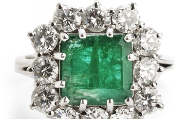 An emerald and diamond ring set with an emerald-cut emerald encircled by numerous brilliant-cut diamonds, mounted in 18k white gold. G-H/VVS-VS. Circa 1950.