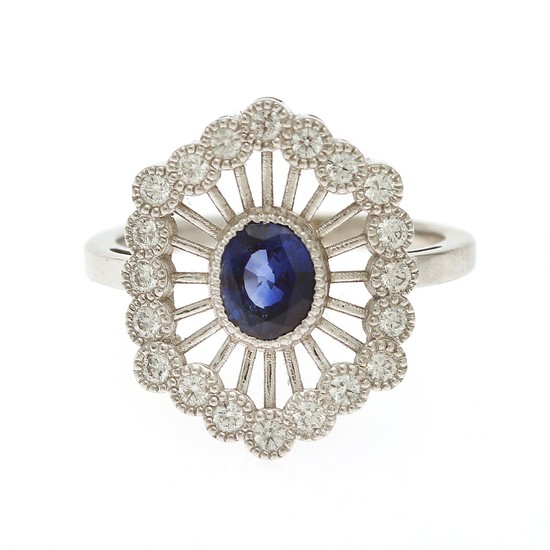A sapphire and diamond ring set with an oval-cut sapphire encircled by numerous brilliant-cut diamonds, mounted in 14k white gold. Size 53.