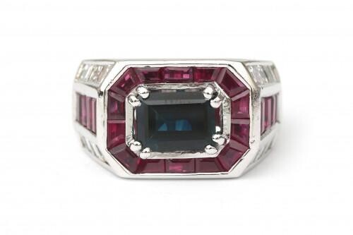 An 18 karat white gold sapphire, ruby and diamond ring. A Of geometrical design featuring a rectangular cut blue sapphire of ca. 2 ct. surrounded by carré cut rubies. The shoulders are set with eight carré cut rubies and sixteen carré cut diamonds, ca...