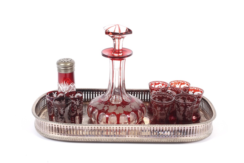 A Bohemian engraved ruby flashed decanter and matched glasses on a tray. The decanter engraved with fruiting vinea, with nine matched engraved shot glasses in differing shapes and a salt shaker similar, the silver-plated shaped rectangular galleried...