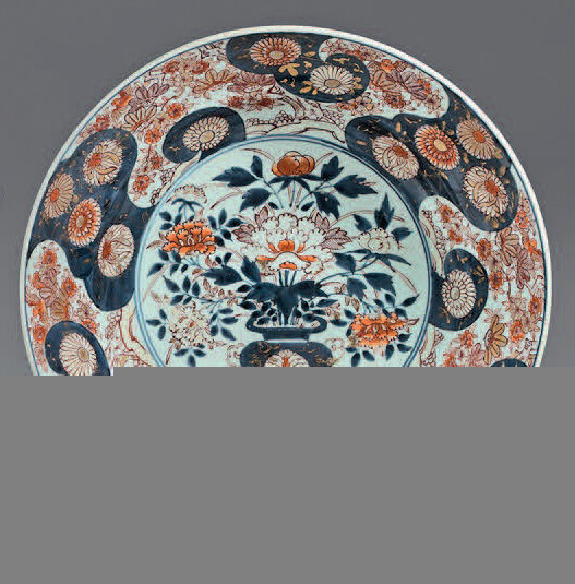 Large Japanese porcelain dish (Arita). 17th century, circa 1680. Imari decoration in the center of a vase of flowers in front of a hedge in a cartouche with two circles, the wing and the fall of flowers in spiral-shaped reserves on a white or blue...