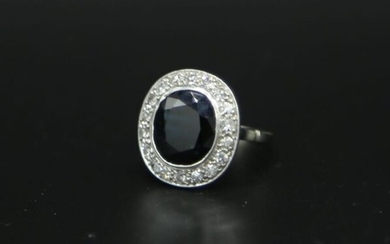Bague en or gris orné d'une pierre bleue ovale entourée de diamants, le panier ajouré de feuillages. Poids brut : 4,79 g / tdd 53 (choc au niveau du serti clos de la pierre centrale)