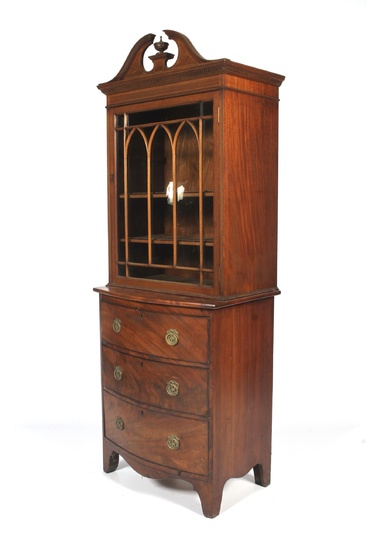 An Edwardian mahogany glazed small bookcase on chest. With broken swan neck pediment and urn finial, with dentil cornice and arched astragal glazed door enclosing two shelves, above three bow fronted drawers with gilt metal wreath handles, on splayed...