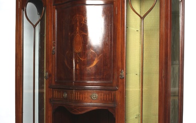 An Edwardian mahogany marquetry inlaid glazed display cabinet. The central cupboard of serpentine form inlaid with neo-classical swags and medallions, enclosing a shelf with lower drawer, flanked by two long elongated glazed display cabinets inlaid...