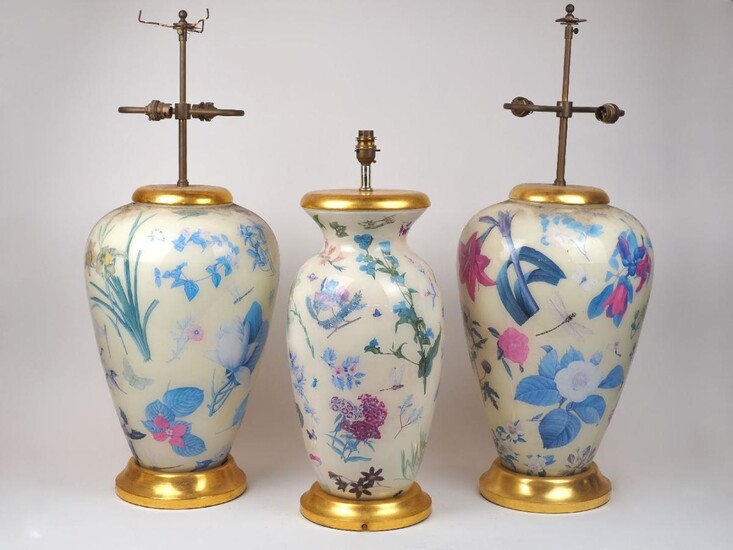 A pair of modern reverse-glass printed botanical lamp bases, decorated with blossoming flowers and insects on a white ground in the decalcomania manner, on gilt bases, 81cm high total each, together with another similar vase, 62cm high (3) It is...