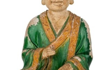A large Chinese fahua-glazed stoneware figure of a monk, Ming dynasty, seated on a square throne in ardha padmasana with right hand raised, head tilted downwards, wearing a voluminous robe covered in green glaze with yellow border decorated in...