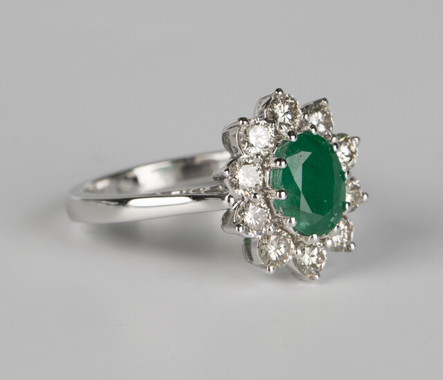 A white gold, emerald and diamond cluster ring, claw set with an oval cut emerald within a surround