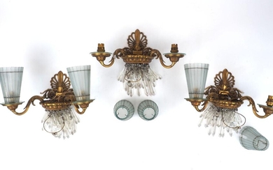 A set of three gilt bronze twin-light wall appliques, 1930s, each with anthemion backplate and foliate scroll branches supporting sectional tapering glass shades, 35cm wide (3) It is the buyer's responsibility to ensure that electrical items are...