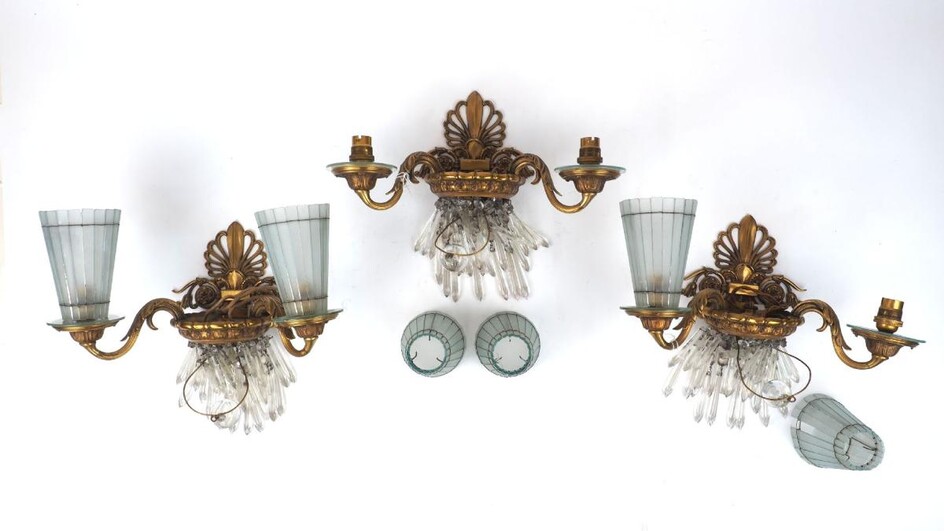A set of three gilt bronze twin-light wall appliques, 1930s, each with anthemion backplate and foliate scroll branches supporting sectional tapering glass shades, 35cm wide (3) It is the buyer's responsibility to ensure that electrical items are...