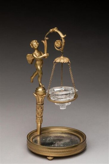 Gilded bronze VEILLEUSE decorated with a cherub standing on a torch, holding in his hands a leafy branch, carrying the crystal tank. Circular base with ice bottom, resting on three runners. Empire period. H. 28, D. 13 cm.