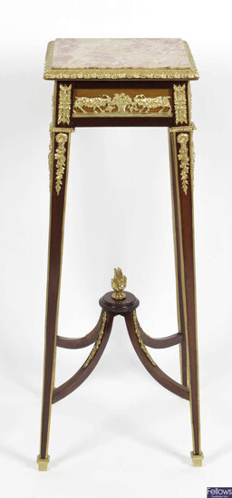 An early 20th century mahogany torchère, the square marble top within a moulded gilt metal border, the frieze with applied gilt metal panels depicting goats with baskets of grapes and vines.